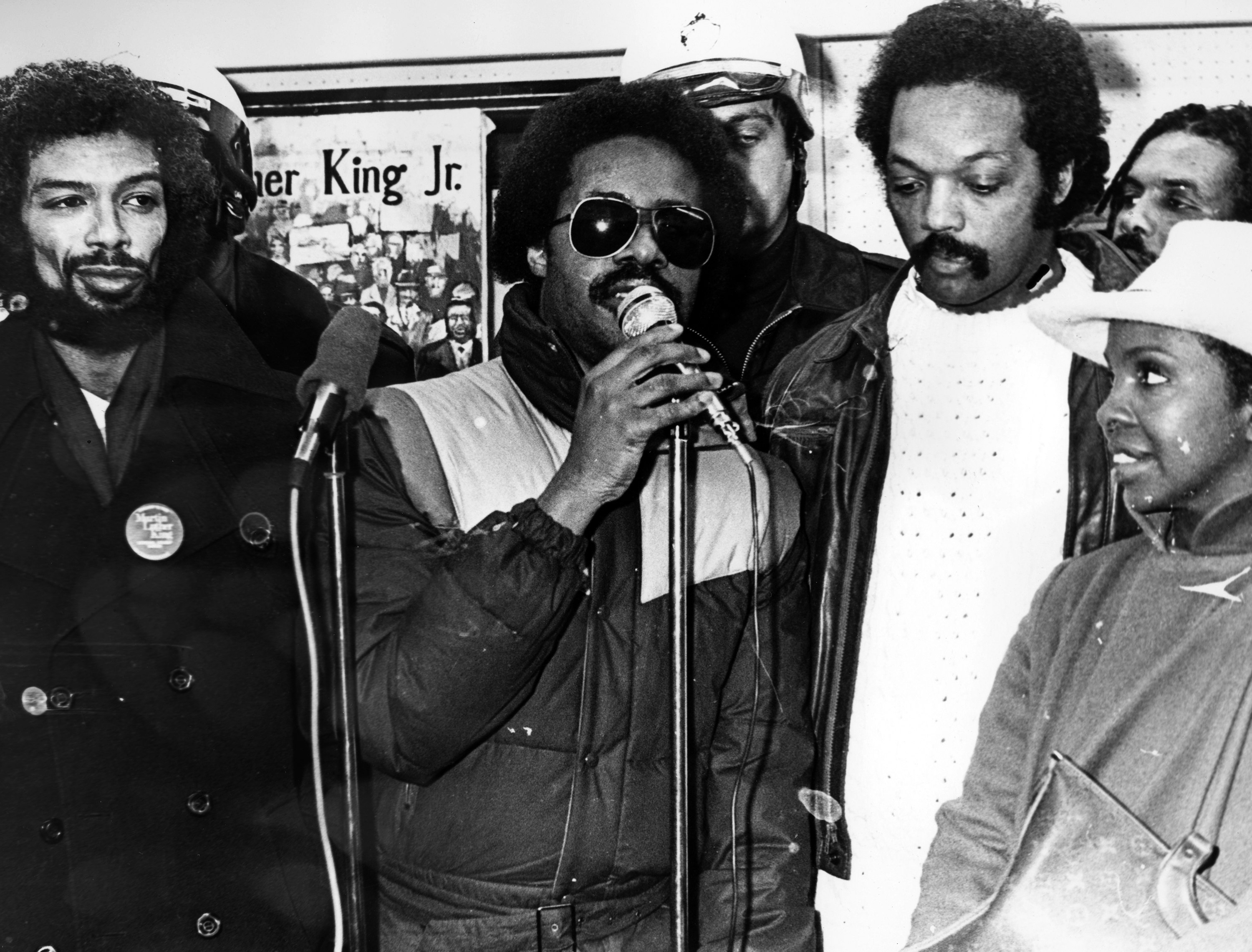 Stevie Wonder, on mic—with Gil Scott-Heron (left), the Reverend Jesse Jackson, singer Gladys Knight, and Sam Courtney—promotes the Martin Luther King Jr. holiday at a press conference at Rafu Gallery, Washington, DC, January 15, 1982. Photo via Afro American Newspapers/Gado/Getty Images.